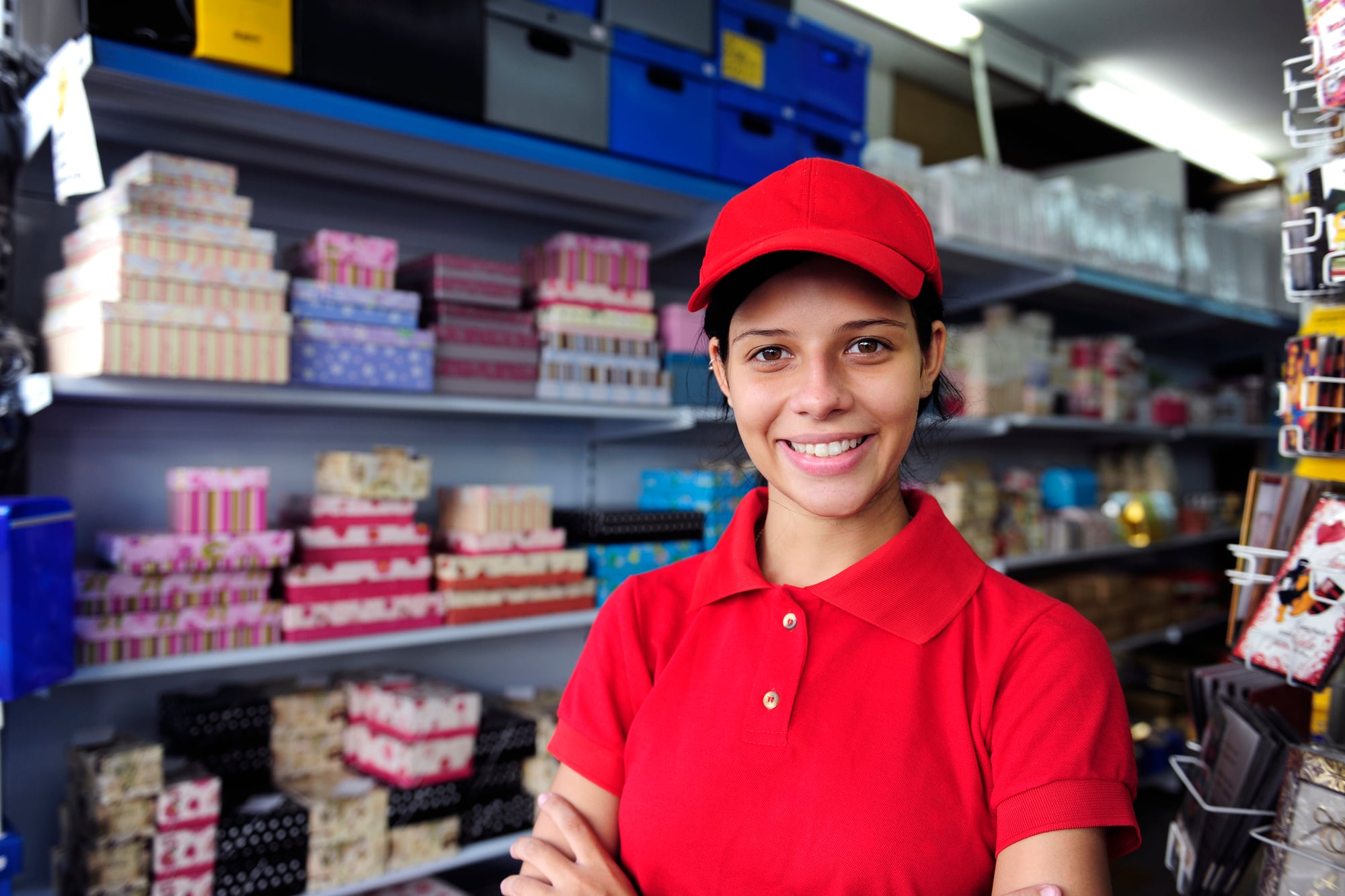 A teen in a red uniform polo and cap standing with arms crossed in a store aisle at their part-time job